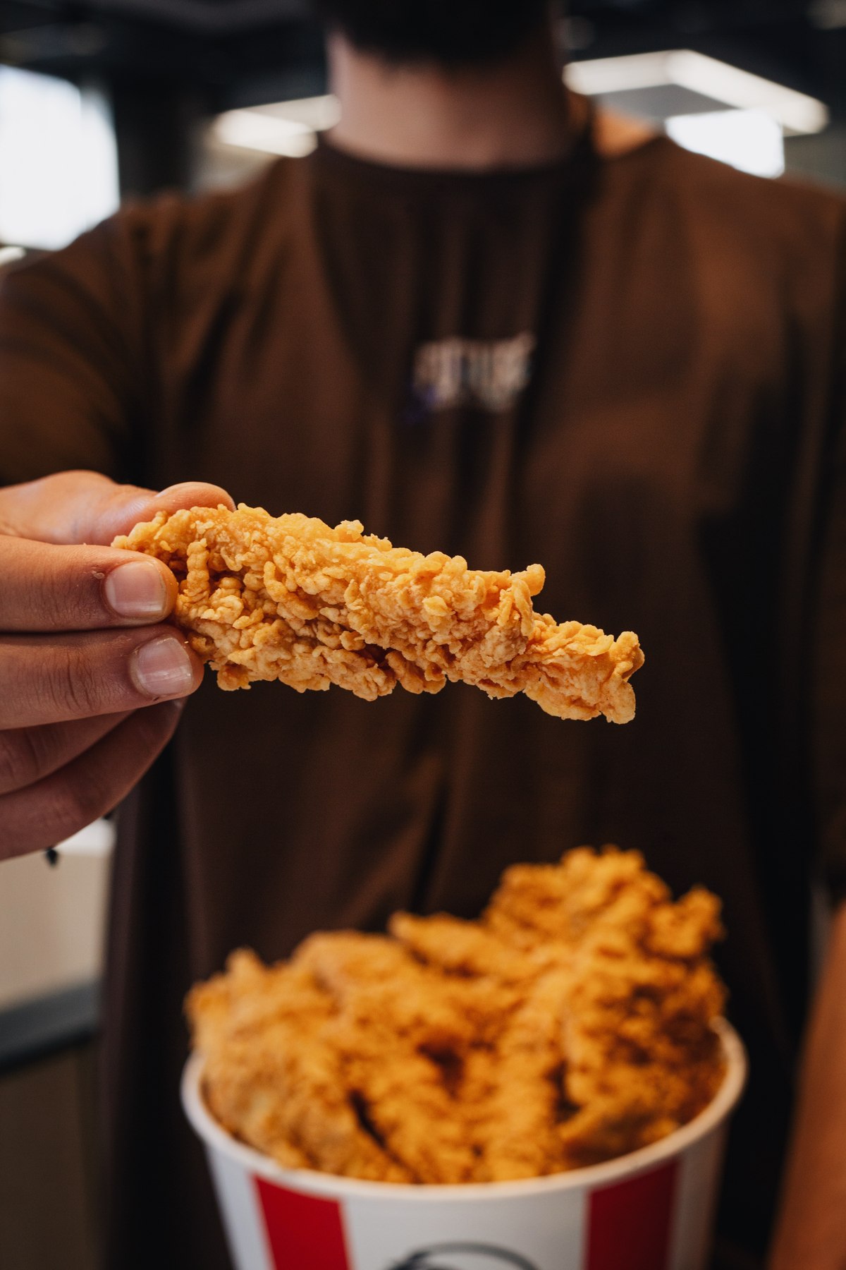 Hand holding a crispy KFC chicken strip above a full bucket
