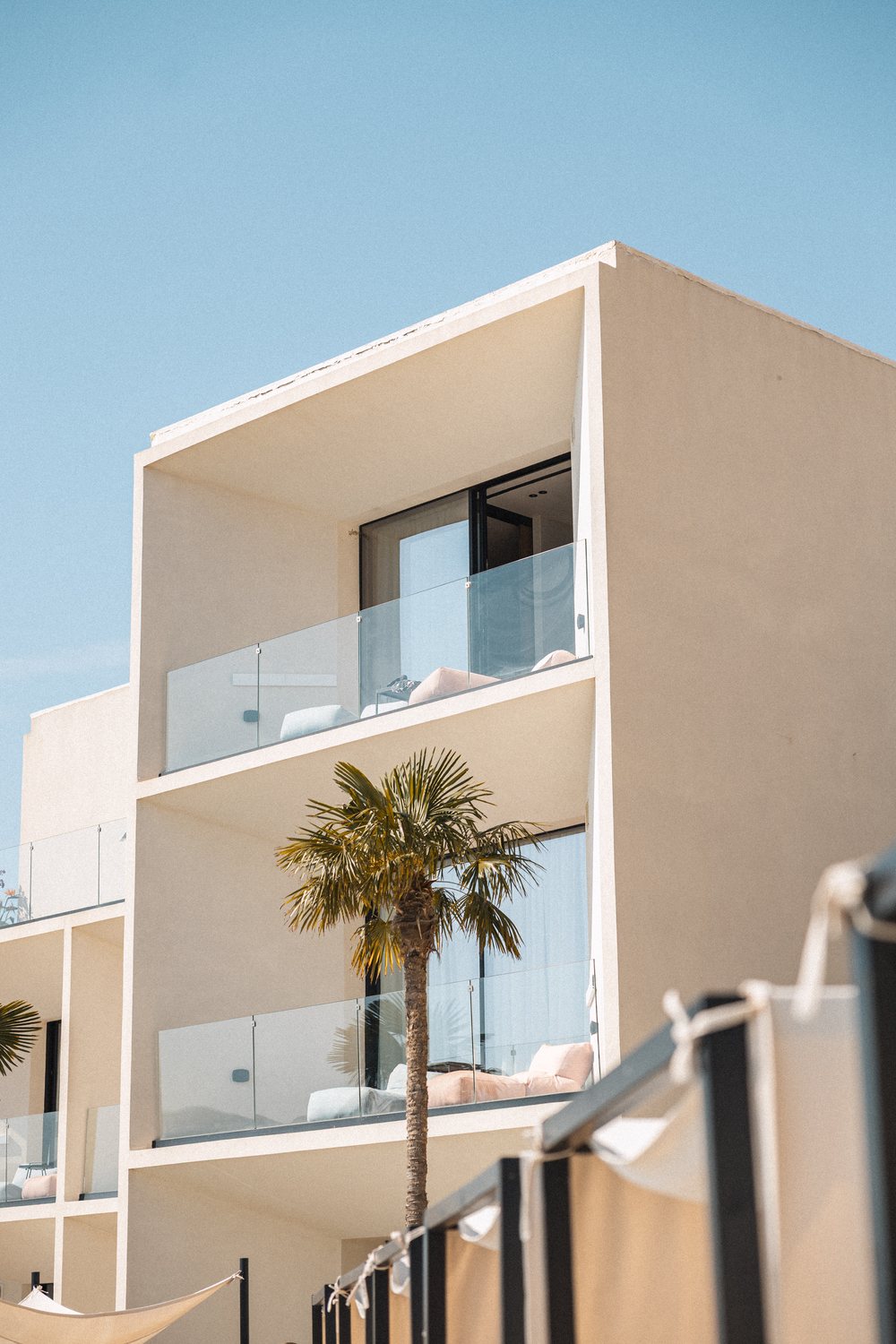 Nobus facade: cream balconies, glass rails and a palm against blue sky