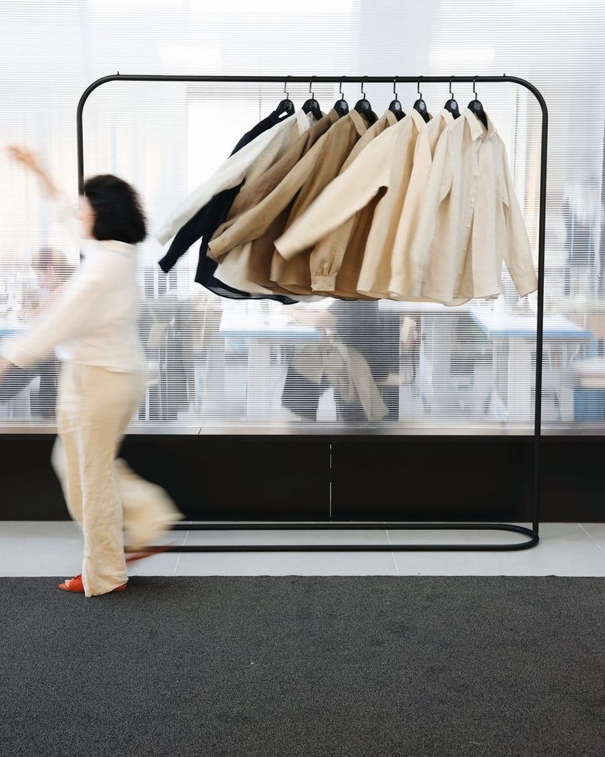 Woman in cream linen moving past a rail of TIERR linen shirts in the atelier