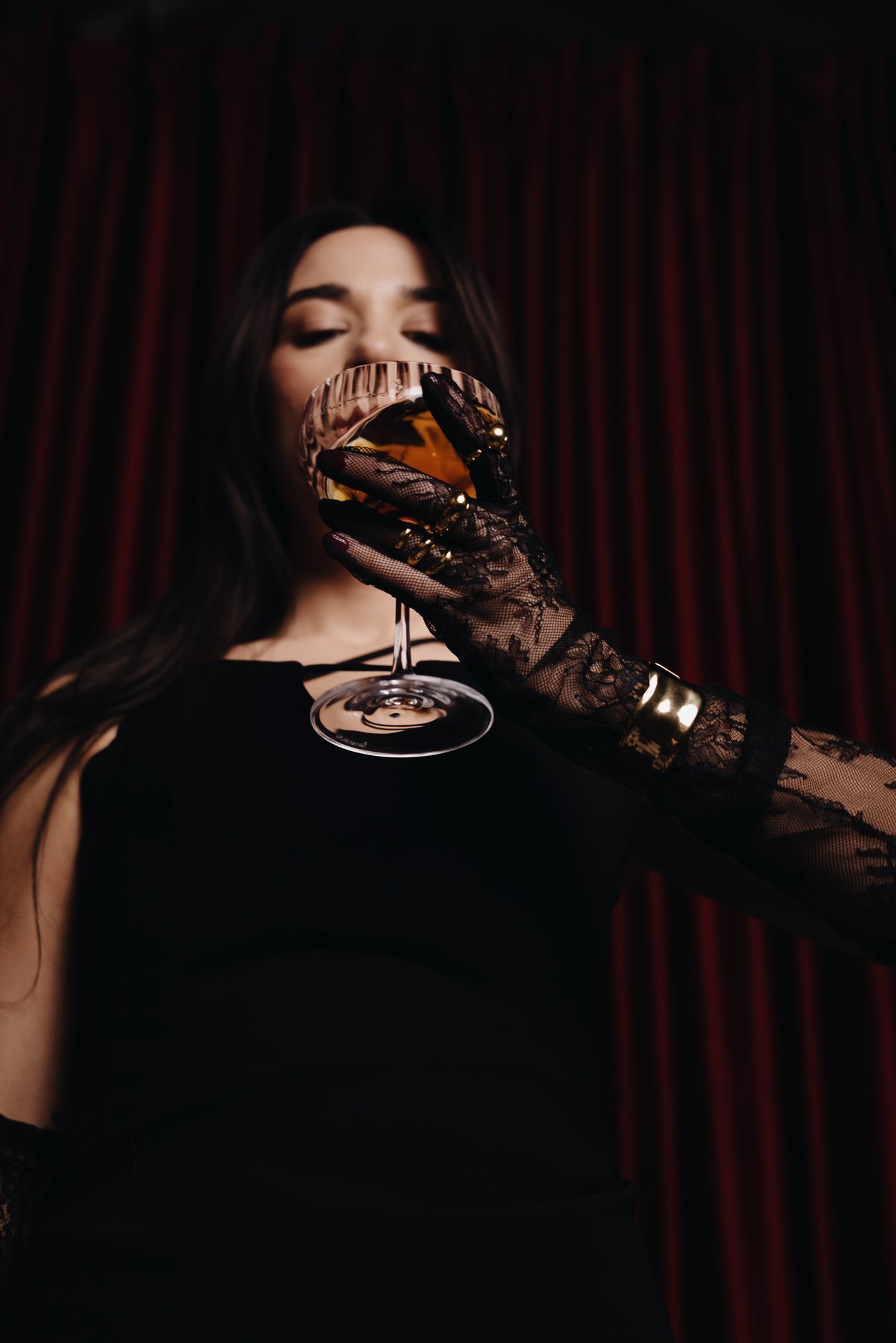 Model sipping from a coupe glass wearing lace gloves, gold rings and cuff