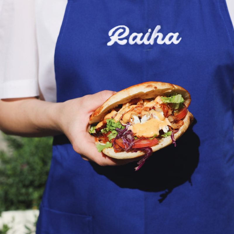 Pita kebab held in front of a blue Raiha apron