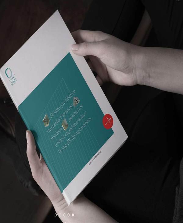The One brochure cover held in hand, teal cover with isometric building illustrations
