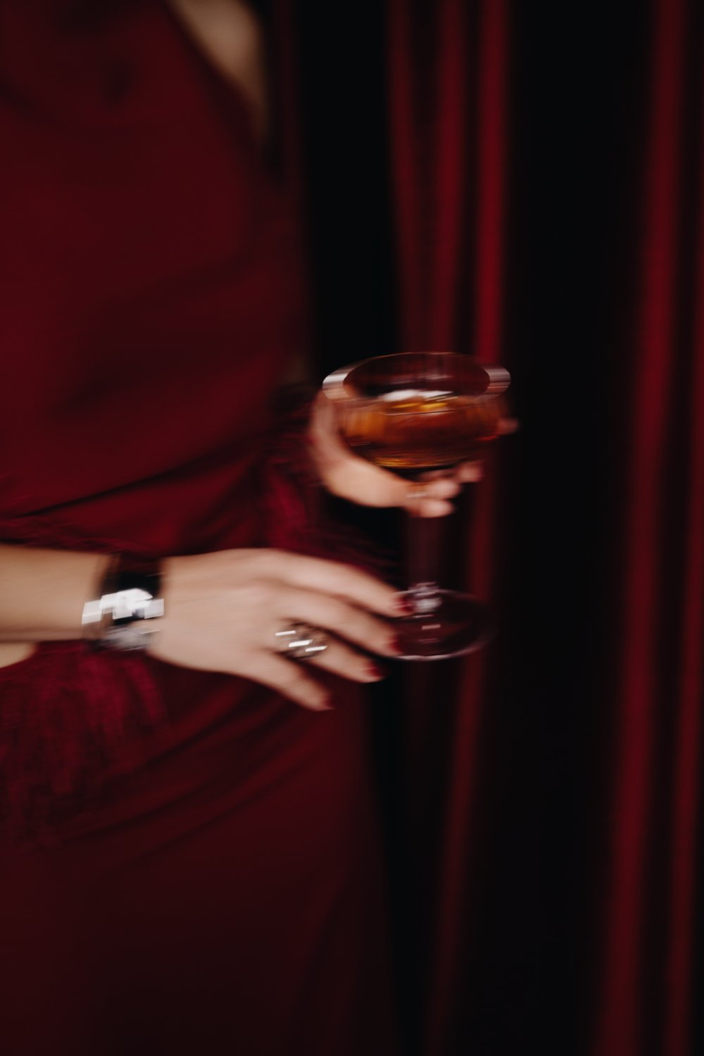 Motion-blurred hand with silver cuff holding a cocktail glass by a red curtain