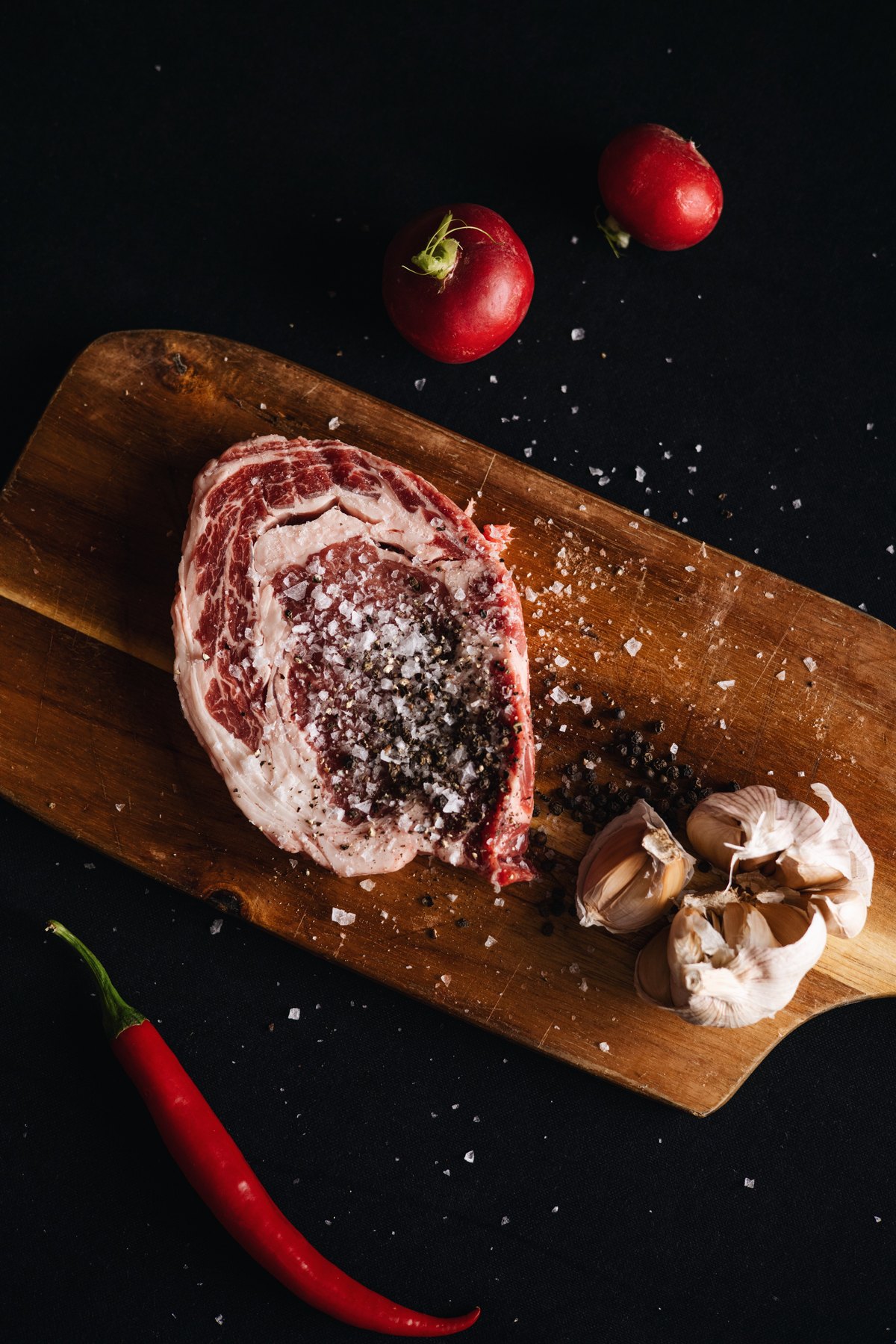 Seasoned ribeye on a wooden board with garlic, radishes and chilli