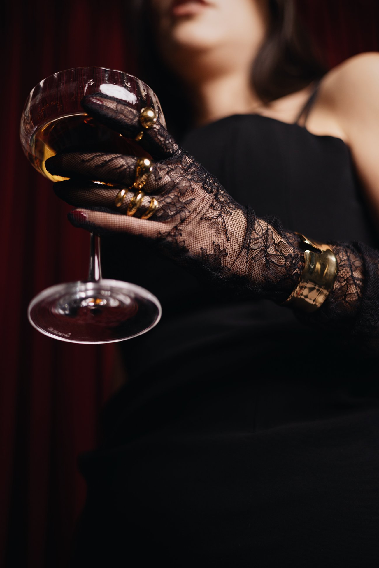 Lace-gloved hand with gold rings and cuff holding a coupe glass