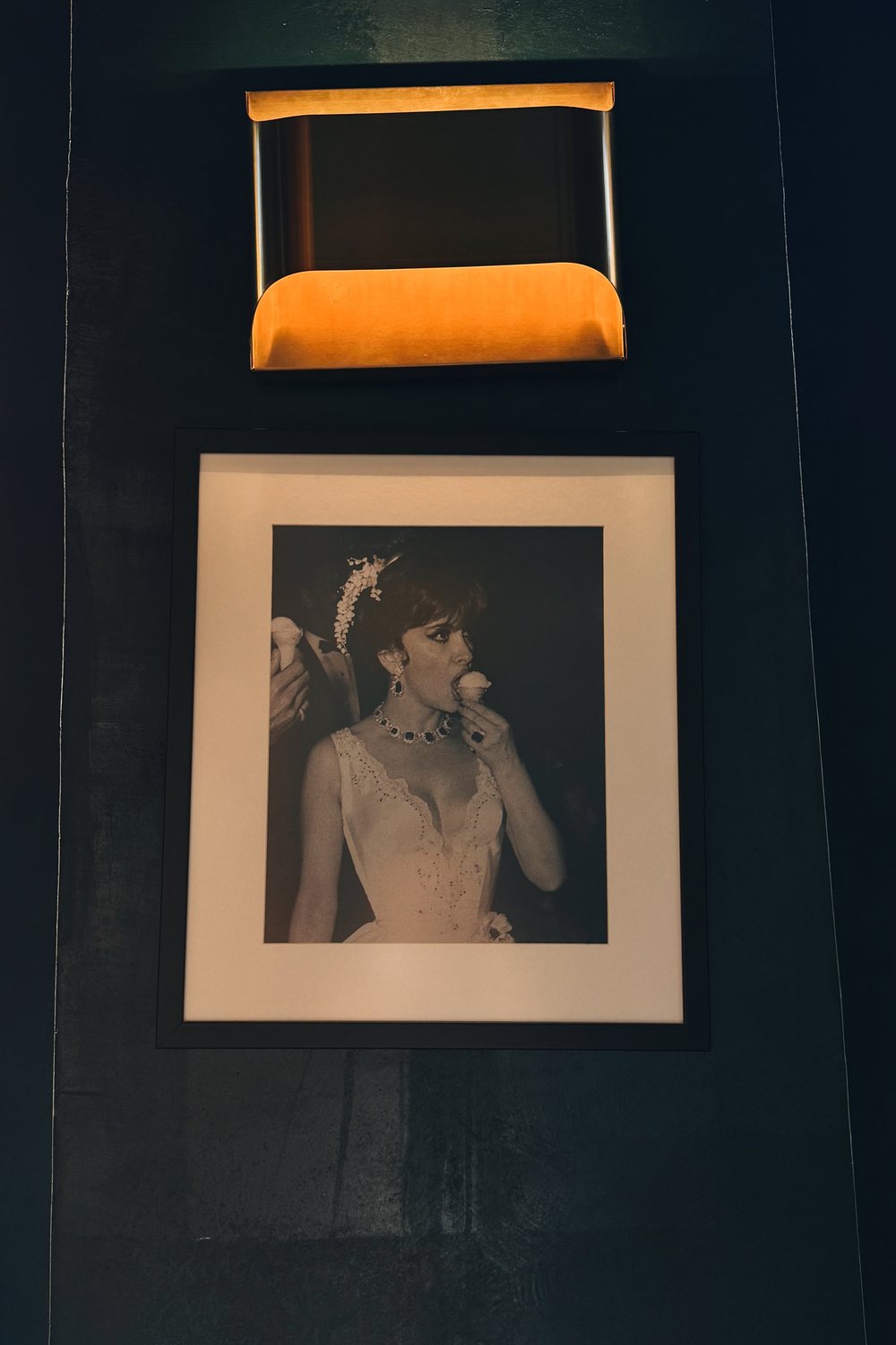 Framed black and white photograph under a brass wall light on a dark green wall