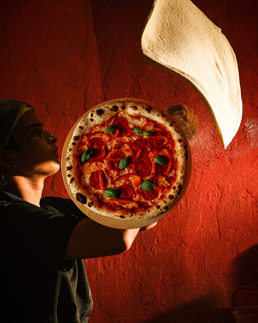 Pizzaiolo holding a pizza with dough flying against a red wall