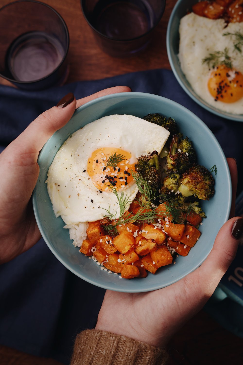 Hands holding a rice bowl with a fried egg, broccoli and sweet potato