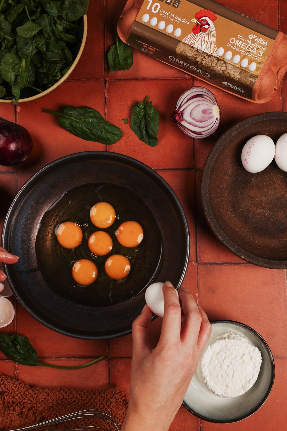 Hand cracking AIBA eggs into a dark pan beside spinach and an Omega 3 carton