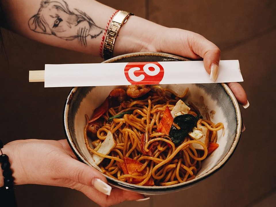 Hands holding a bowl of Sushi Co noodles with branded chopsticks